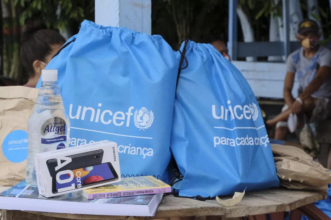 Foto mostra algumas mochilas azuis com o logo do UNICEF em cima de um banco e o conte&uacute;do de um kit conectividade: caderno, livro, celular, &aacute;lcool em gel, m&aacute;scaras
