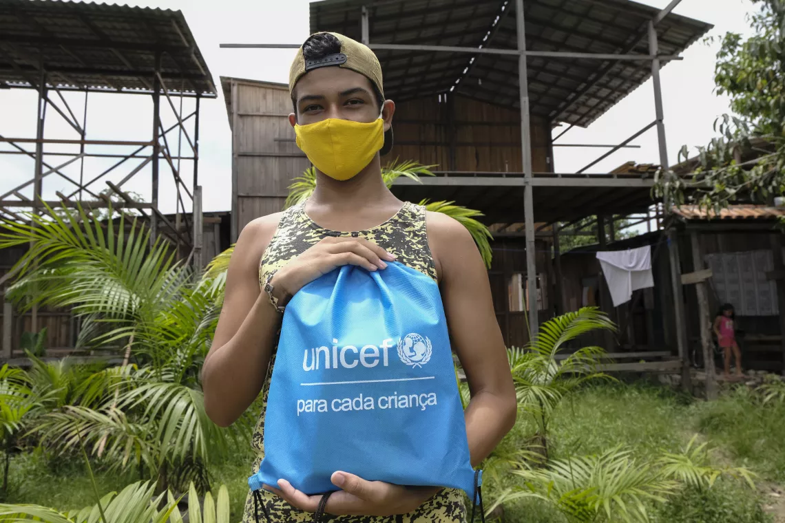 Foto mostra um adolescente em p&eacute; segurando uma mochila com o logo do UNICEF. Ele est&aacute; de m&aacute;scara e usa bon&eacute; com a aba virada para tr&aacute;s. O adolescente est&aacute; em frente a uma constru&ccedil;&atilde;o de madeira. H&aacute; tamb&eacute;m muita vegeta&ccedil;&atilde;o em volta dele.