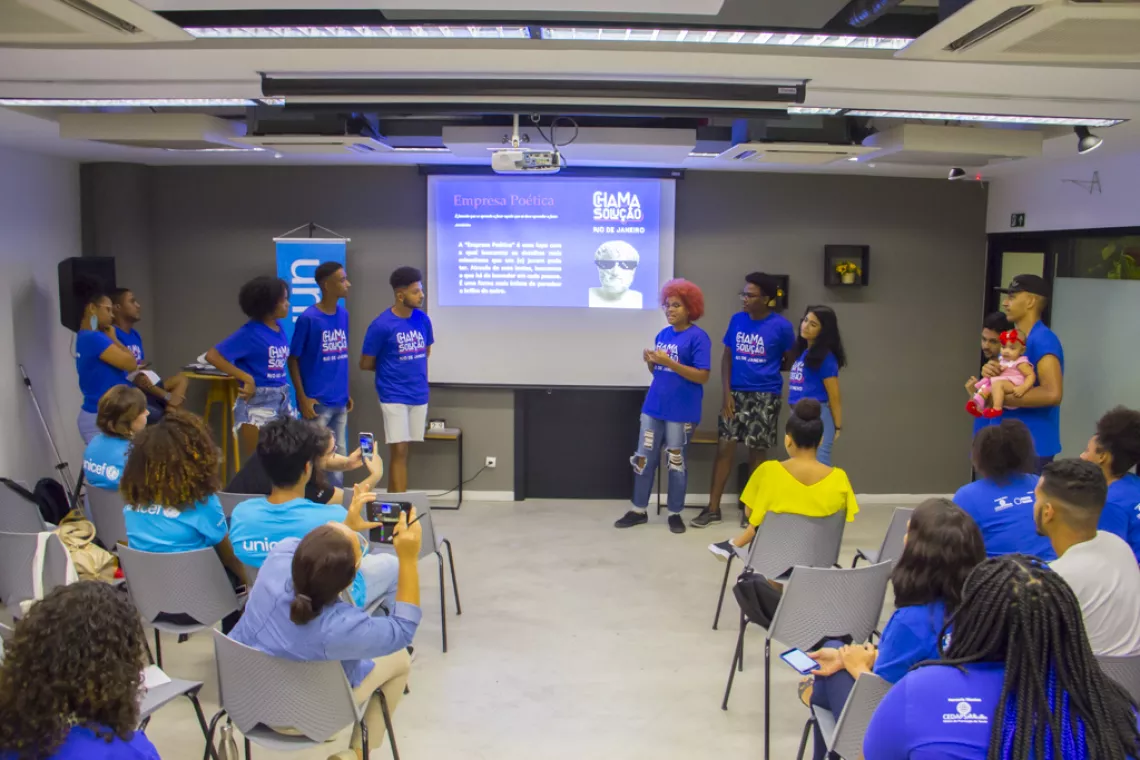 foto de pessoas fazendo uma apresenta&ccedil;&atilde;o de powerpoint, seis pessoas est&atilde;o em p&eacute; e outras sentadas em cadeiras enfileiradas.