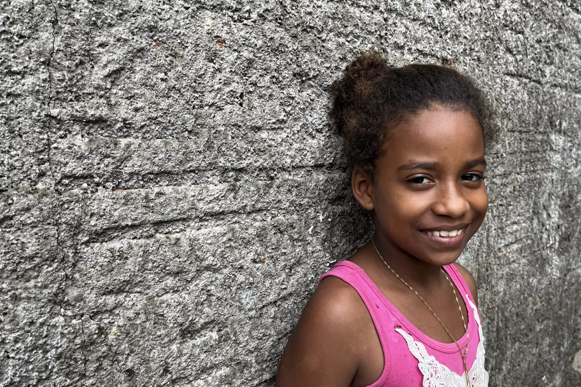 Foto mostra uma menina encostada em uma parede de adobe. Ela est&aacute; olhando para a c&acirc;mera e sorrindo.