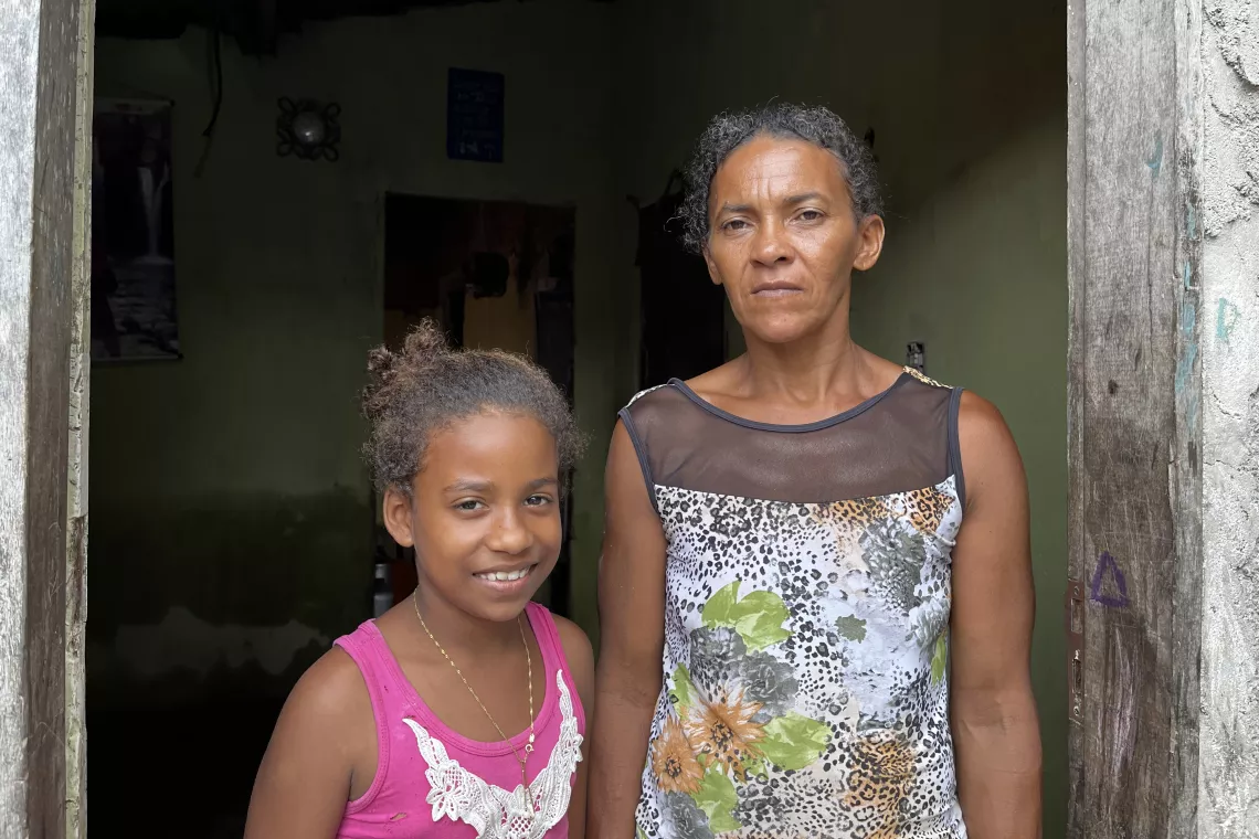 Foto mostra uma menina do lado de sua m&atilde;e. As duas est&atilde;o olhando para a foto. A menina est&aacute; sorrindo. A m&atilde;e est&aacute; s&eacute;ria.