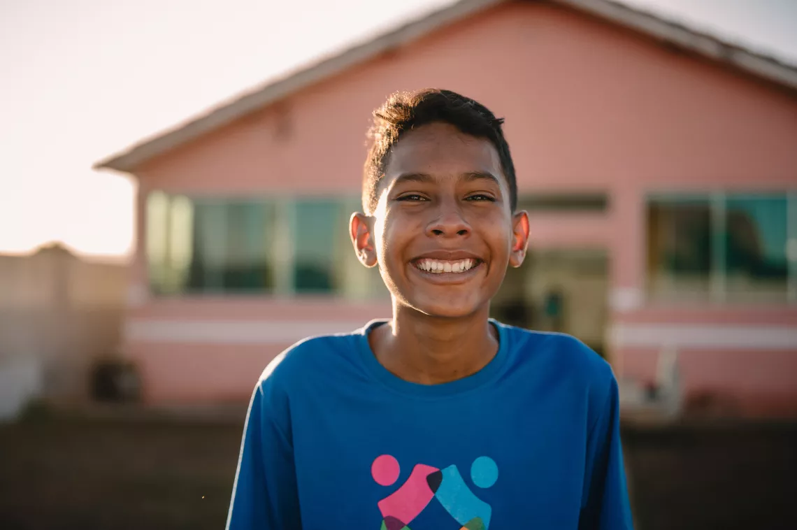 Foto mostra um adolescente sorridente. Ele est&aacute; ao ar livre em frente a uma casa rosada.