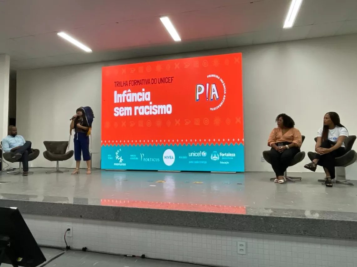 Foto mostra um palco com algumas pessoas sentadas e uma adolescente em p&eacute; falando no microfone.