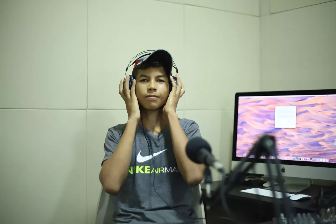 foto mostra um adolescente sentado em um estúdio de rádio, com as mãos no fone de ouvido