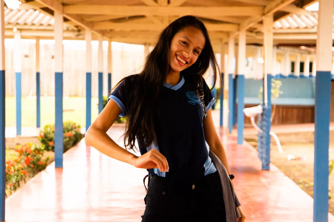 Foto mostra uma adolescente sorridente, em p&eacute;, no corredor de uma escola. 