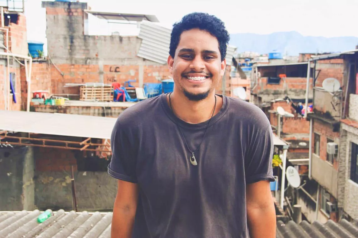 Morador da Mar&eacute;, no Rio de Janeiro, Thiago Carlos participa do projeto #CRIAndoRede, no qual aprendeu o que &eacute; sa&uacute;de mental e como cada um pode se cuidar. Na foto Thiago est&aacute; sorridente apoiado em um muro. Atr&aacute;s dele est&atilde;o casas da comunidade onde mora