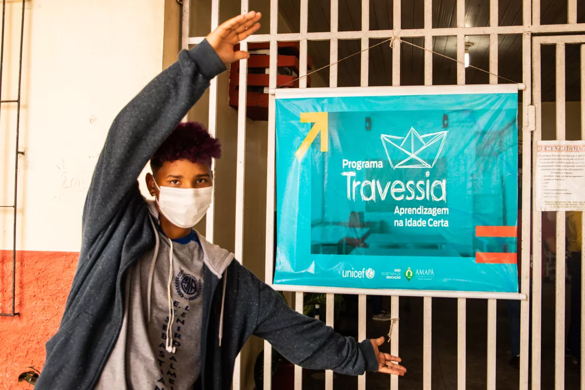 Foto mostra um adolescente em frente a uma grade onde est&aacute; pendurado um banner do projeto Travessia.