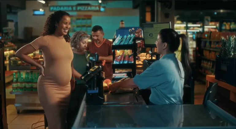 imagem retirada de uma campanha sobre parto que mostra mulher gr&aacute;vida conversando com caixa de supermercado. ao fundo, uma senhora e um homem na fila