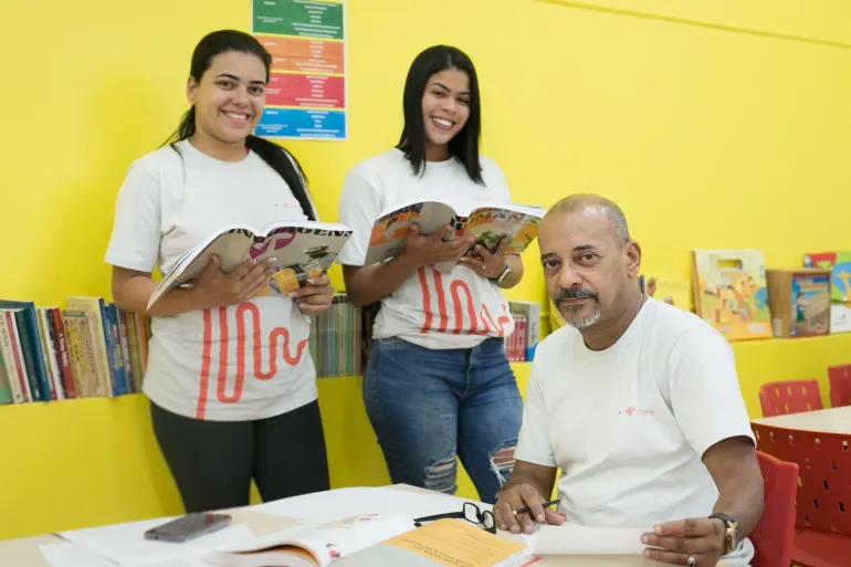 duas mulheres e um home posam para foto em uma sala de aula com parede amarela