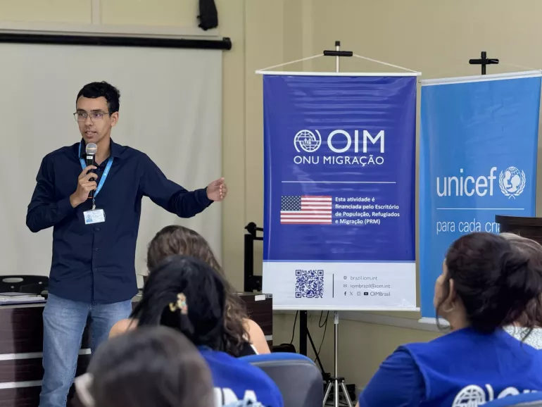 Homem vestindo camisa azul e crach&aacute; ministra palestra. Ao fundo, banners da OIM e do UNICEF
