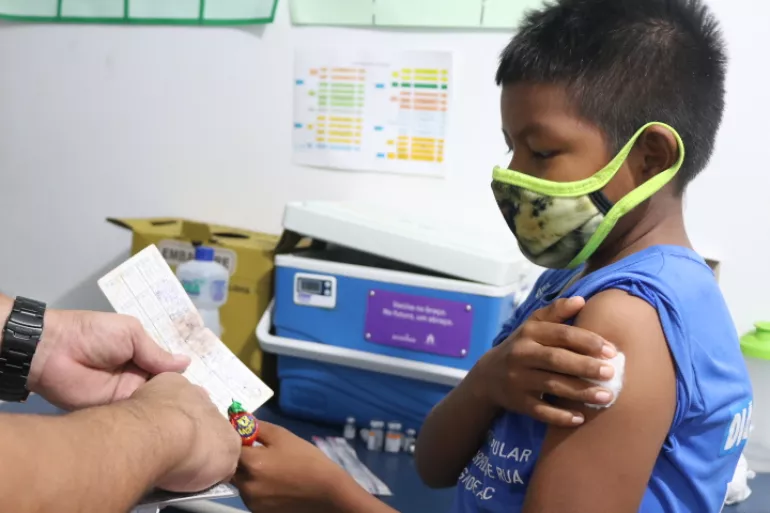 Foto mostra um menino de máscara segurando um algodão no braço depois de receber uma vacina. Ele está de perfil. Com a outra mão, ele segura a caderneta de vacinação, para onde está olhando.