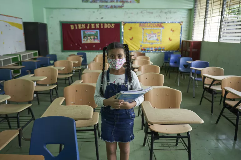uma menina usando m&aacute;scara e segurando material escolar est&aacute; parada em p&eacute; no meio de uma sala de aula vazia