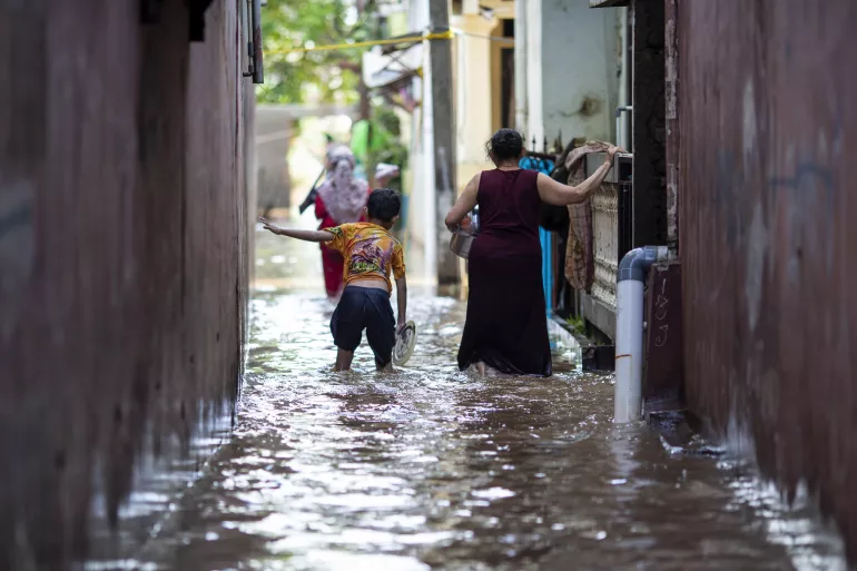 Em 21 de fevereiro de 2021, um menino e sua m&atilde;e caminham atrav&eacute;s das &aacute;guas em uma &aacute;rea afetada por enchentes no leste de Jacarta, Indon&eacute;sia.