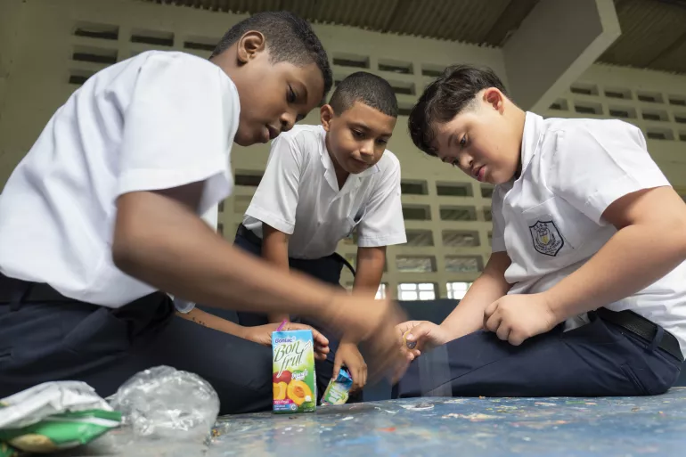 Foto mostra tr&ecirc;s adolescentes sentados no ch&atilde;o do p&aacute;tio da escola. Um dos meninos tem s&iacute;ndrome de Down.