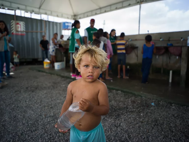 um menino pequeno, sem camisa, segurando uma garrafa pl&aacute;stica amassada, olha para a c&acirc;mera, s&eacute;rio. Ele est&aacute; em uma barraca de acampamento para migrantes. Ao fundo da foto, h&aacute; outras pessoas.