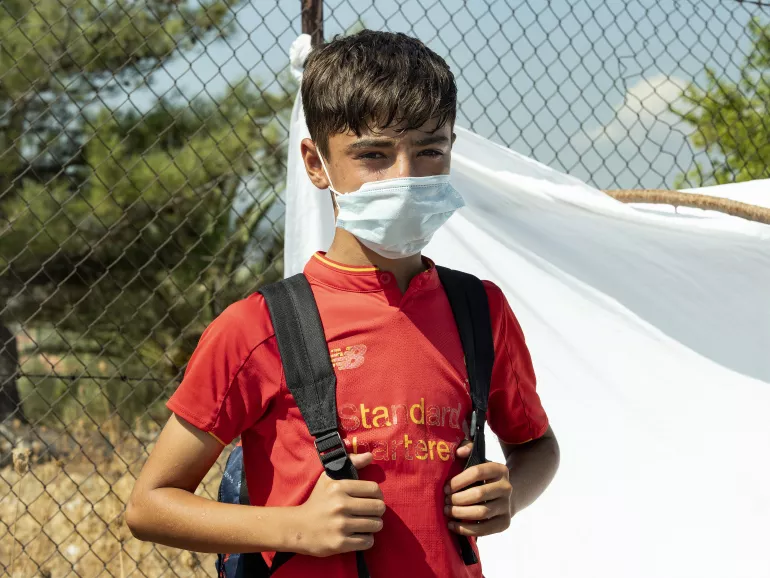 Um menino usando m&aacute;scara e carregando uma mochila olha para a c&acirc;mera. Ele est&aacute; em p&eacute; em um espa&ccedil;o aberto e atr&aacute;s dele h&aacute; um len&ccedil;ol branco.