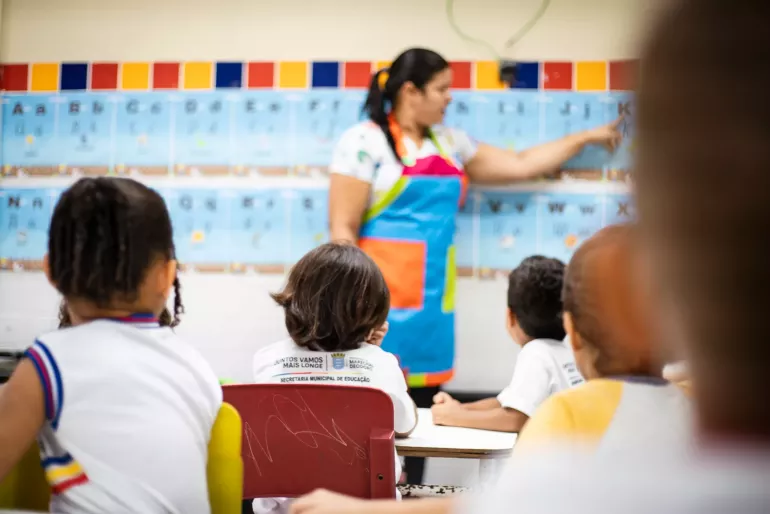Sala de aula onde professora mostra algo no quadro e crian&ccedil;as aparecem de costas assistindo &agrave; aula