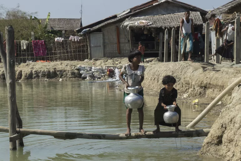 Duas meninas pequenas buscam &aacute;gua em um a&ccedil;ude no campo de Nget Chaung, em Rakhine, Mianmar. Elas est&atilde;o sobre um tronco fino de madeira e parecem n&atilde;o aguentar o peso dos jarros.