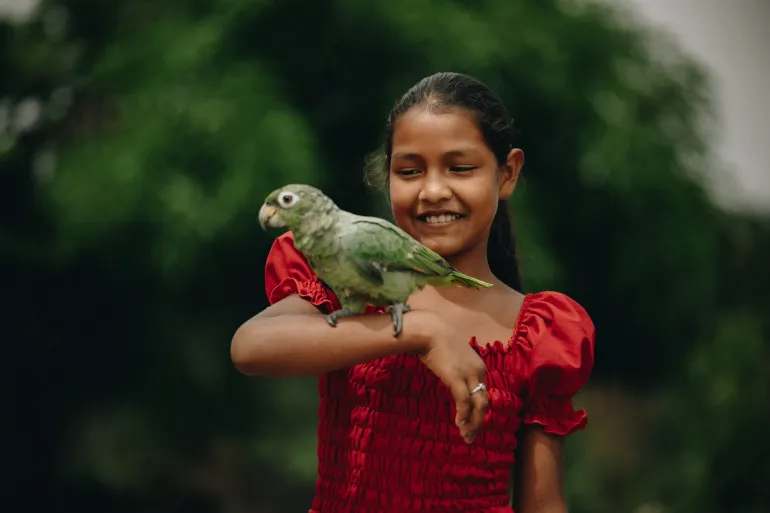 menina de vestido vermelho segura um papagaio verde na m&atilde;o direita e sorri para a foto