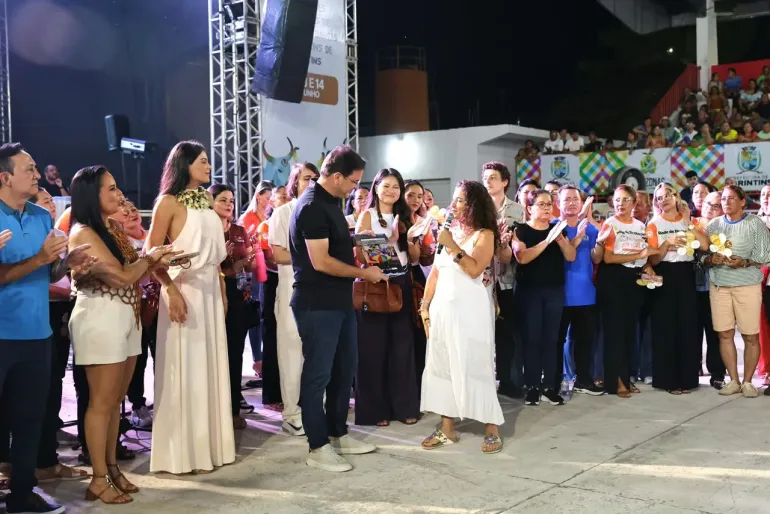 grupo de adultos em p&eacute; enquanto uma mulher de branco fala ao microfone