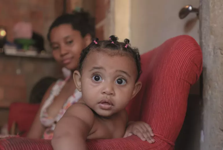 M&atilde;e e menina olham para a c&acirc;mera sentadas em um sof&aacute; vermelho 