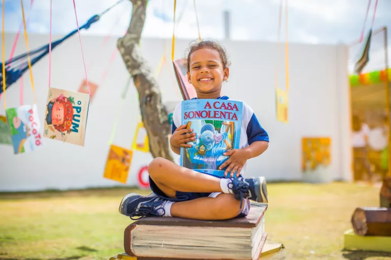 uma menina est&aacute; sentada em um banco em formato de pilha de livros. ela segura um livro nas m&atilde;os, em frente ao peito.
