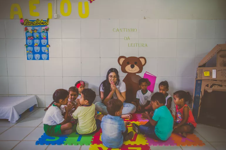 Um grupo de crian&ccedil;as pequenas est&atilde;o em volta da educadora que est&aacute; com as m&atilde;os juntas, contando uma hist&oacute;ria