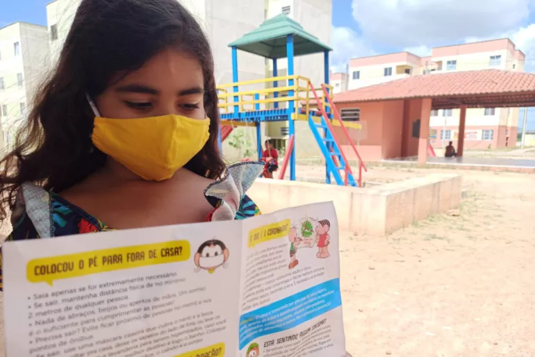menina usando m&aacute;scara l&ecirc; um folheto com personagens da Turma da M&ocirc;nica, atr&aacute;s dela um conjunto habitacional e um parque infantil