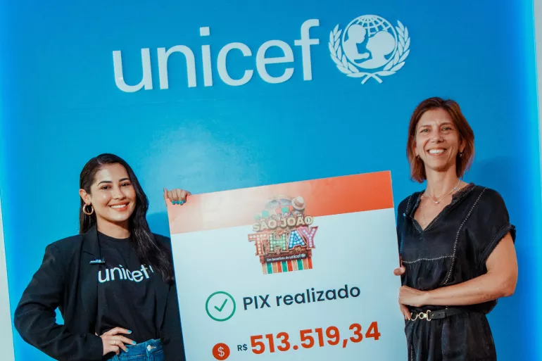 Foto mostra Thaynara OG, vestindo a camiseta do UNICEF, segurando um banner junto com a representante interina do UNICEF no Brasil, Paola Babos. As duas est&atilde;o em frente a uma parede azul com o logo do UNICEF. Elas est&atilde;o sorrindo.