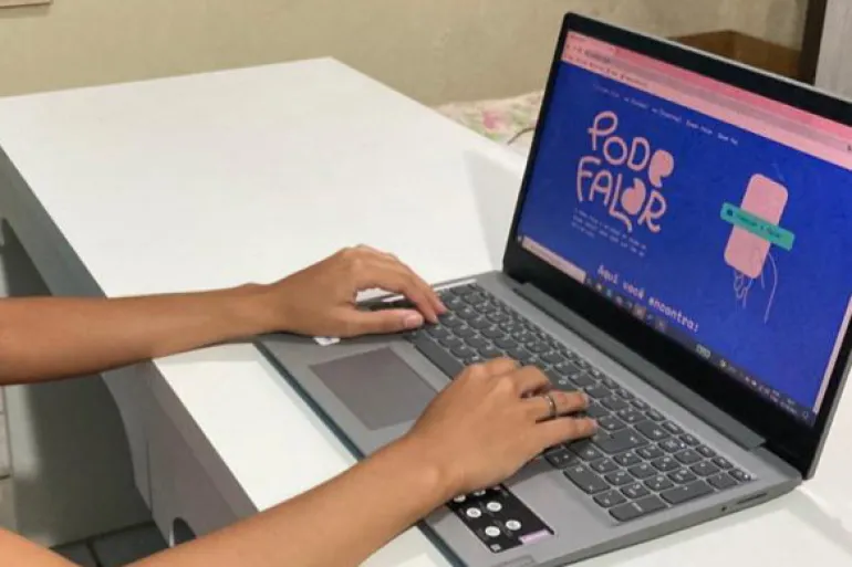 Foto mostra as m&atilde;os de uma mulher em cima de um teclado de laptop. Na tela do teclado, a janela est&aacute; aberta no site do Pode Falar.