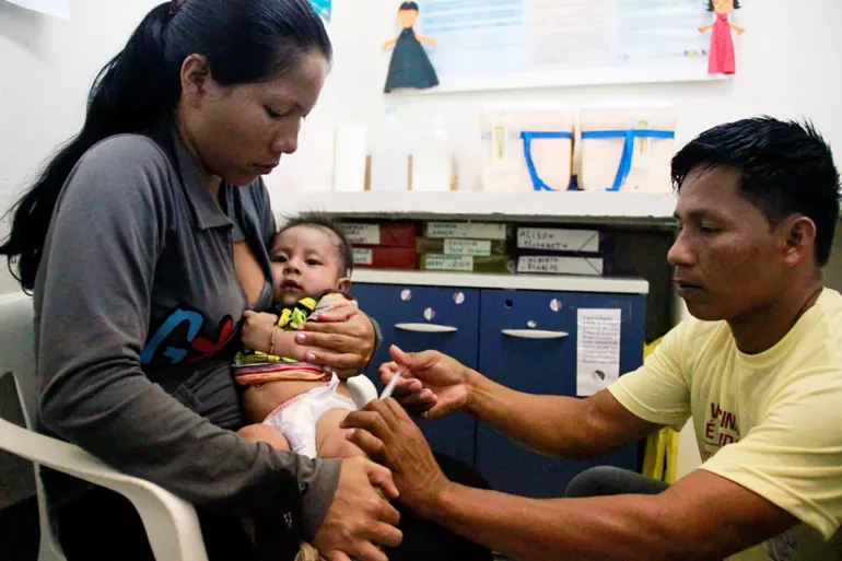 Foto mostra um beb&ecirc; ind&iacute;gena sendo vacinado. Ele est&aacute; no colo da m&atilde;e. O profissional de sa&uacute;de est&aacute; agachado na frente deles.