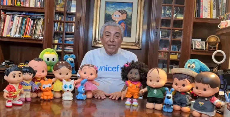O escritor Mauricio de Sousa est&aacute; sentado atr&aacute;s de uma mesa onde est&atilde;o bonecos de muitos dos personagens criados por ele.