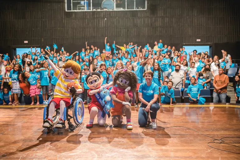 Foto mostra um audit&oacute;rio com crian&ccedil;as e adultos na plateia e no palco os personagens Luca, M&ocirc;nica e Milena, al&eacute;m de a chefe do escrit&oacute;rio do UNICEF em S&atilde;o Paulo
