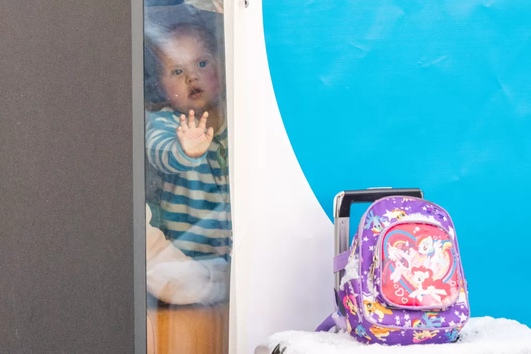 Foto mostra um beb&ecirc; olhando atrav&eacute;s de uma parede de vidro. Ao lado dele, h&aacute; um c&iacute;rculo azul e uma mochila infantil.