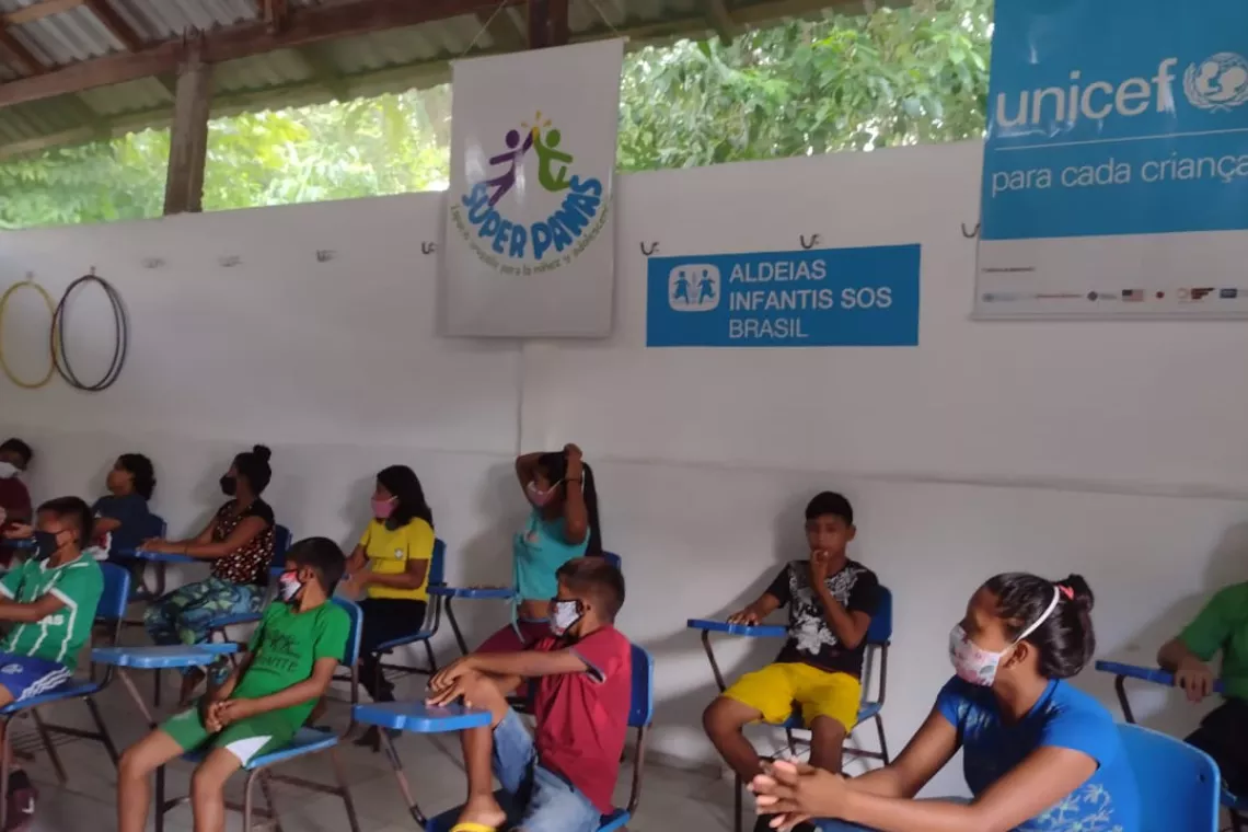 v&aacute;rios adolescentes e crian&ccedil;as est&atilde;o sentados em carteiras escolares olhando para algu&eacute;m que n&atilde;o aparece na foto. todos usam m&aacute;scaras