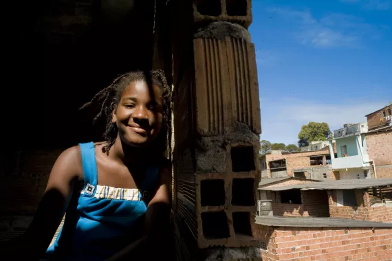 Adolescente est&aacute; na janela de casa, atr&aacute;s dela casas de tijolo aparente de uma comunidade de Salvador.