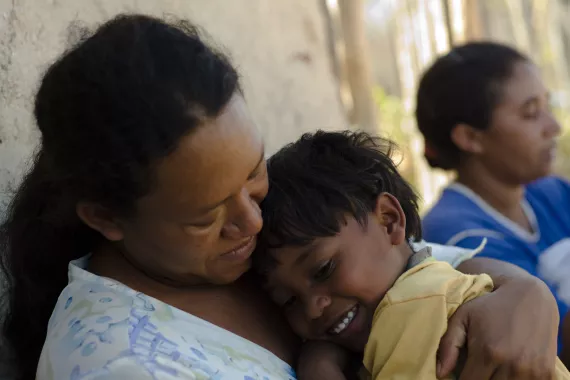 Um menininho ind&iacute;gena est&aacute; no colo de sua m&atilde;e, que o abra&ccedil;a.