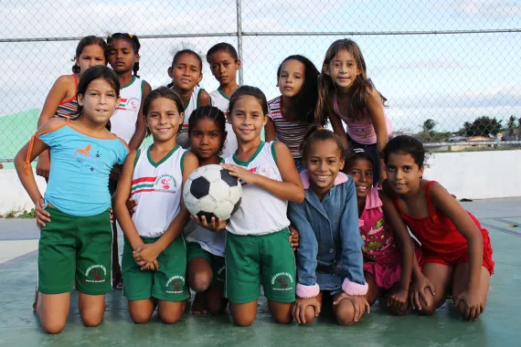 um grupo de meninas est&aacute; numa quadra de esportes. Algumas est&atilde;o ajoelhadas na frente de outras. A menina no centro segura uma bola.