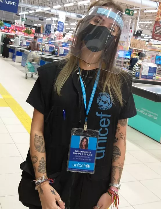 uma mulher est&aacute; em p&eacute; dentro de um supermercado; ela usa m&aacute;scara, escudo facial, colete do UNICEF e crach&aacute;