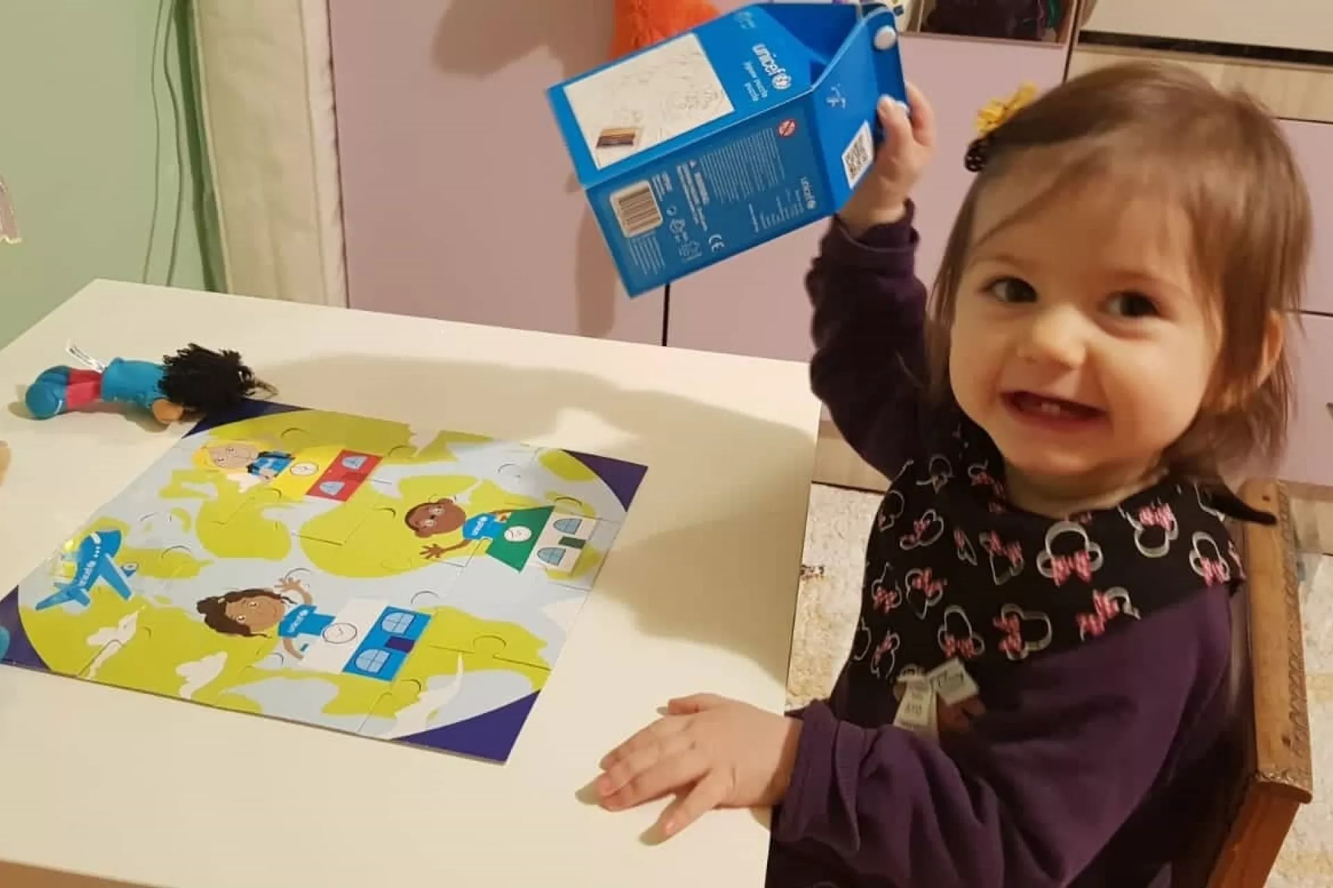 Малко момиченце си играе в стаята си с пъзел, гледа към камерата и се усмихва