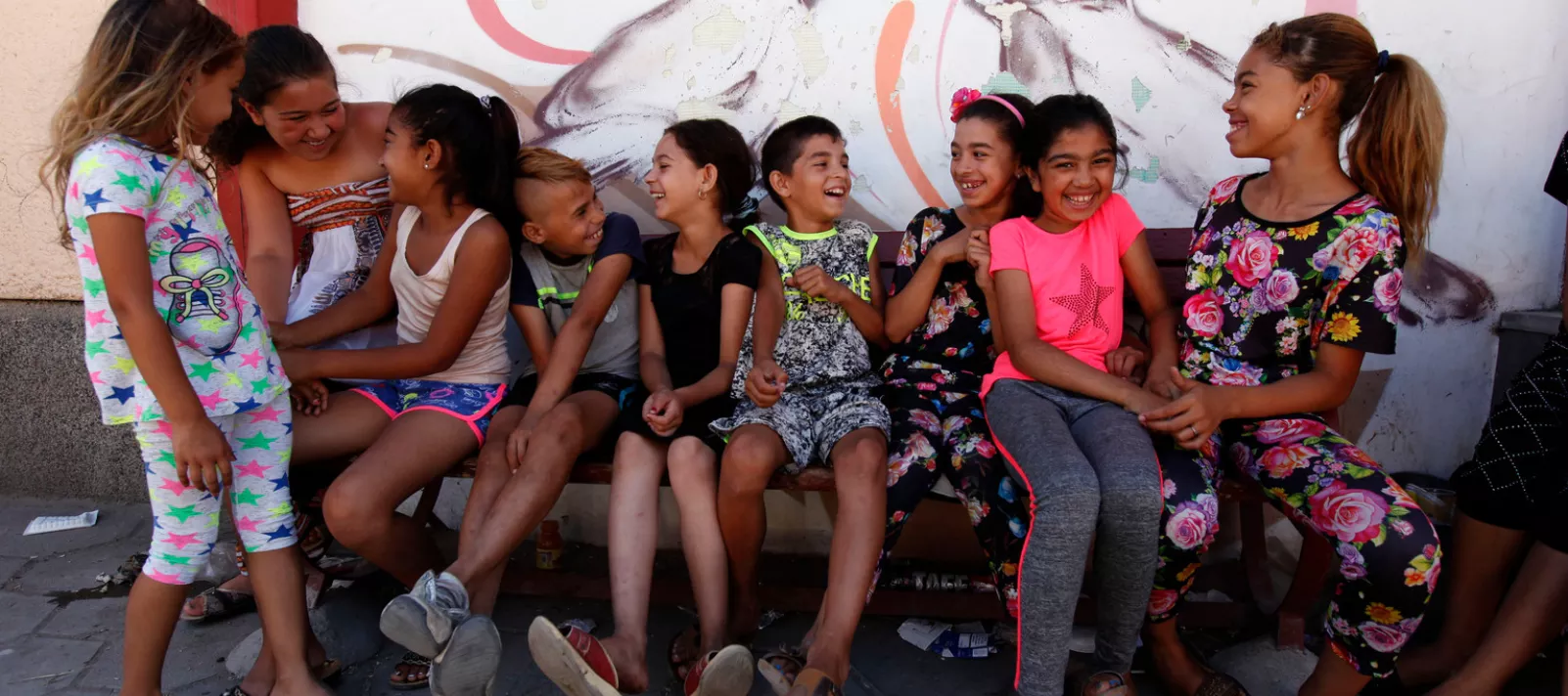 Деца от уязвими общности са седнали на пейка и се забавляват, зад тях има красиви графити