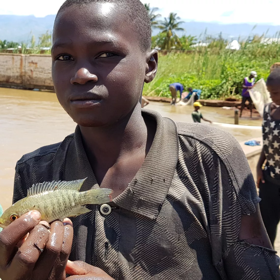 Jean-Pierre holds the catch of the day from the water puddle in the background. Jean-Pierre does not go to school anymore because his school is closed.