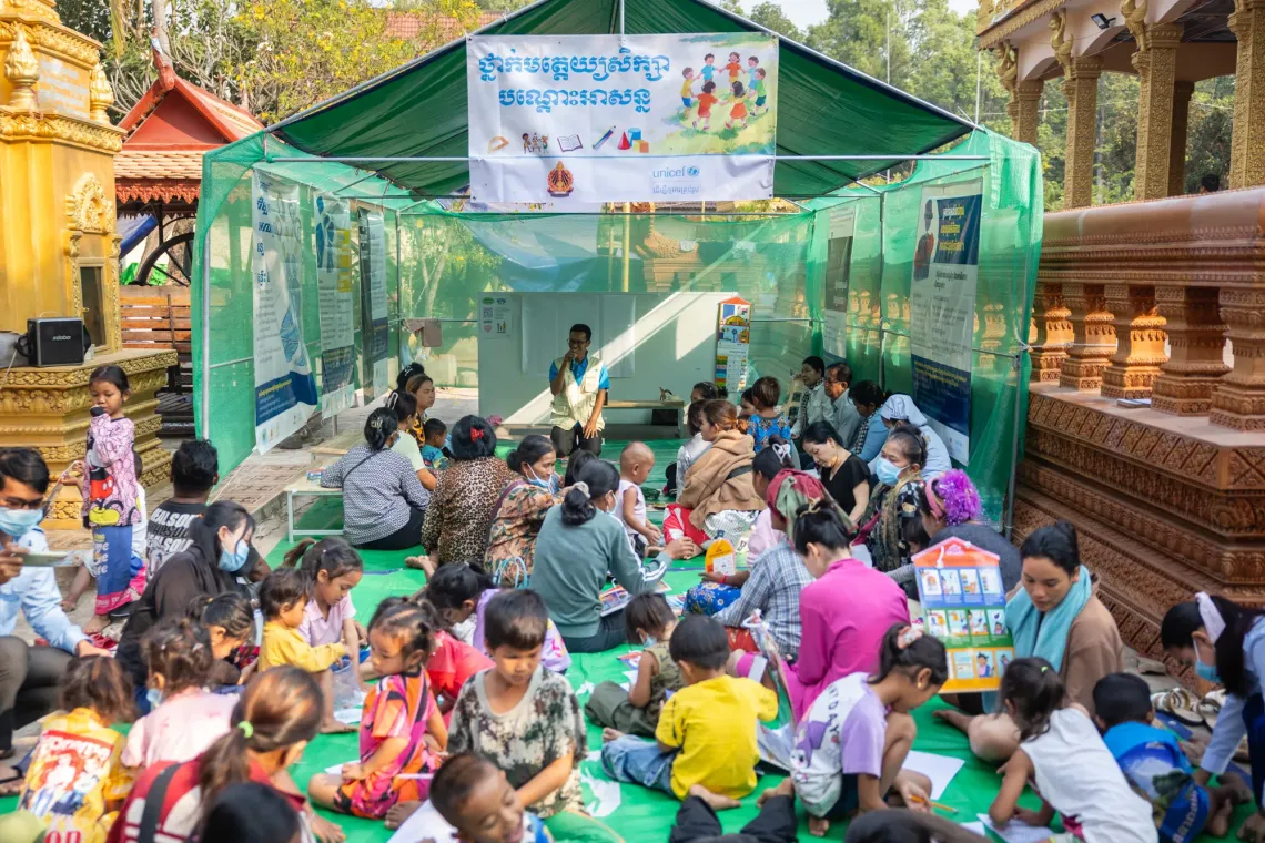 A young boy draws while his parent listens to a parenting session nearby supported by MoEYS and UNICEF.