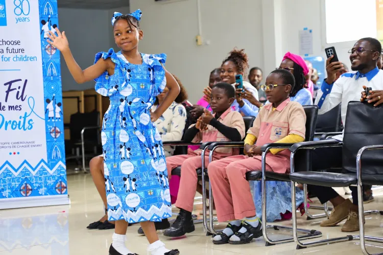 A child models a dress made from UNICEF Cameroon&rsquo;s 80th anniversary commemorative fabric, &ldquo;The Thread of My Rights,&rdquo; during its unveiling event in Yaound&eacute;. Audience members, including children and adults, watch the presentation while a UNICEF80 display stands in the background.