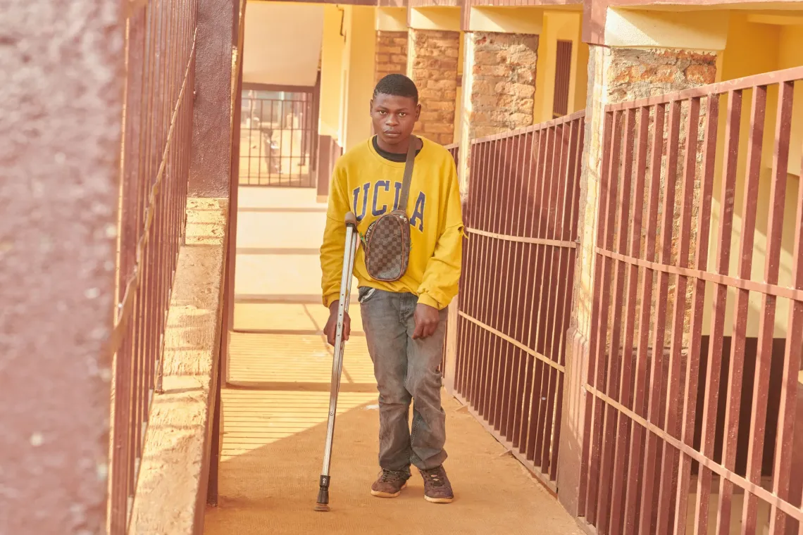 A teenage boy with a physical disability walks across a school compound using crutches, smiling as he heads toward his classroom.
