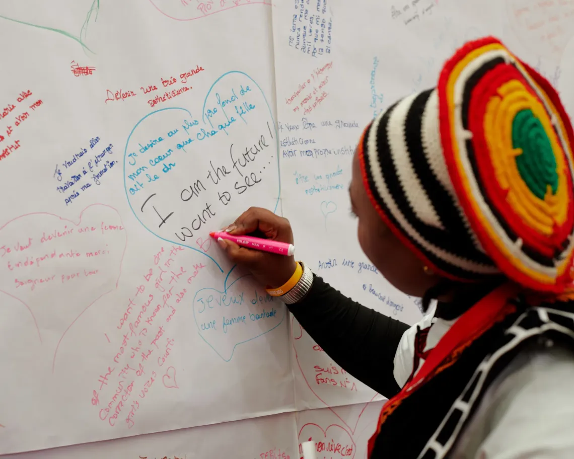 Person wearing a striped hat writes a message on a large white sheet filled with colorful handwritten notes and hearts.
