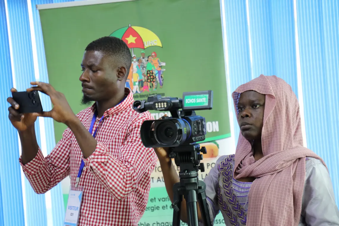 Two media professionals record content during a health communication event, with one using a smartphone and the other operating a video camera mounted on a tripod. A health promotion display is visible in the background.