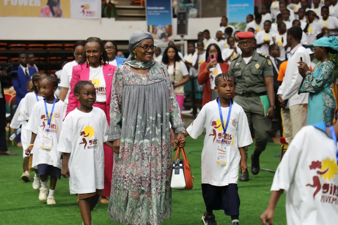 Adult leading children wearing Girls&rsquo; Movement T-shirts during a public event with a large crowd in the background.