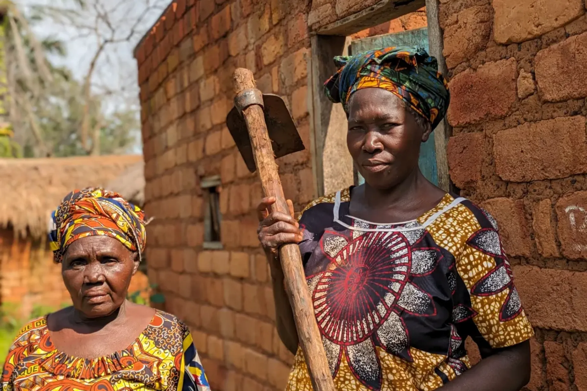 Antoinette Adiambi, hoe in hand, stands for a photo next to her damaged house in Koumboli village. 