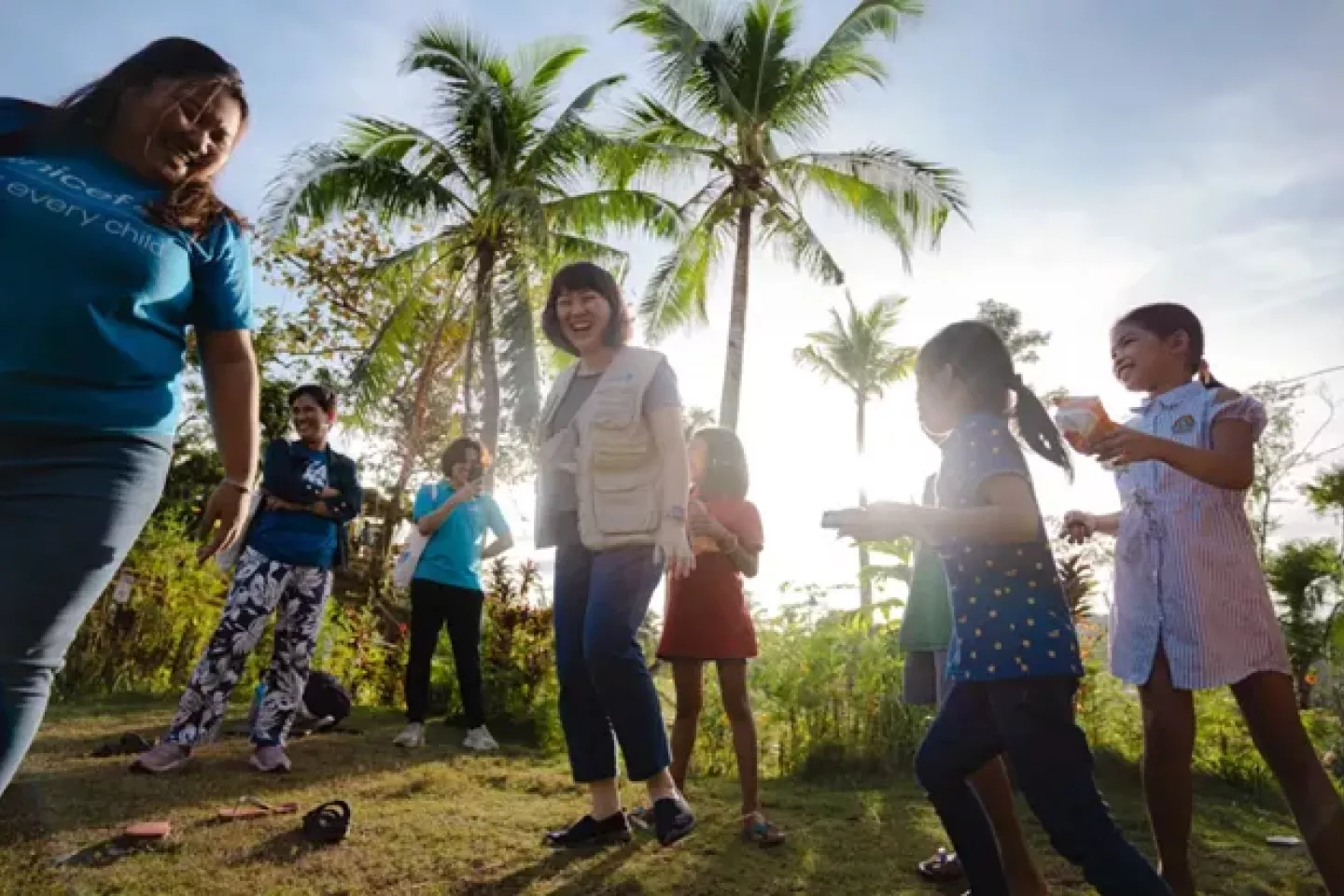 Women and children standing in a forest area and smiling. There are palm trees in the background